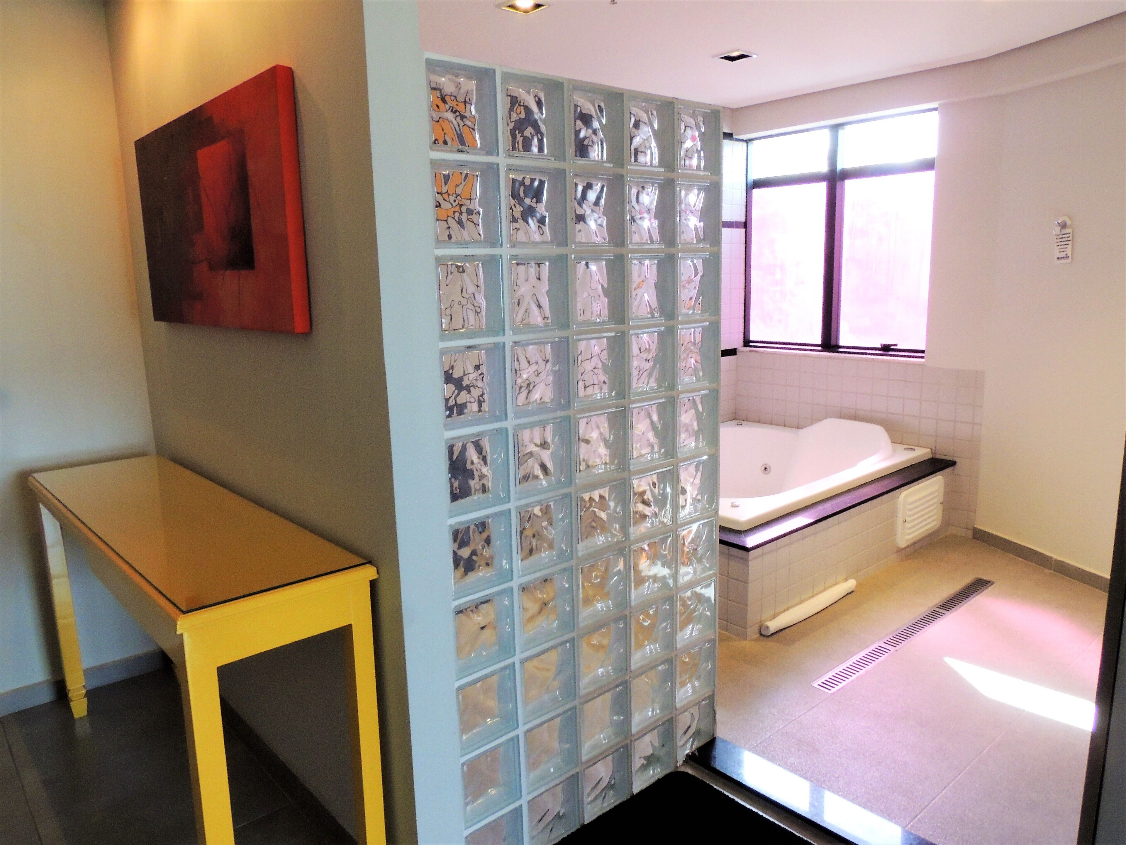 A bathroom with a bathtub, a glass partition with storage, a window, and a yellow table.
