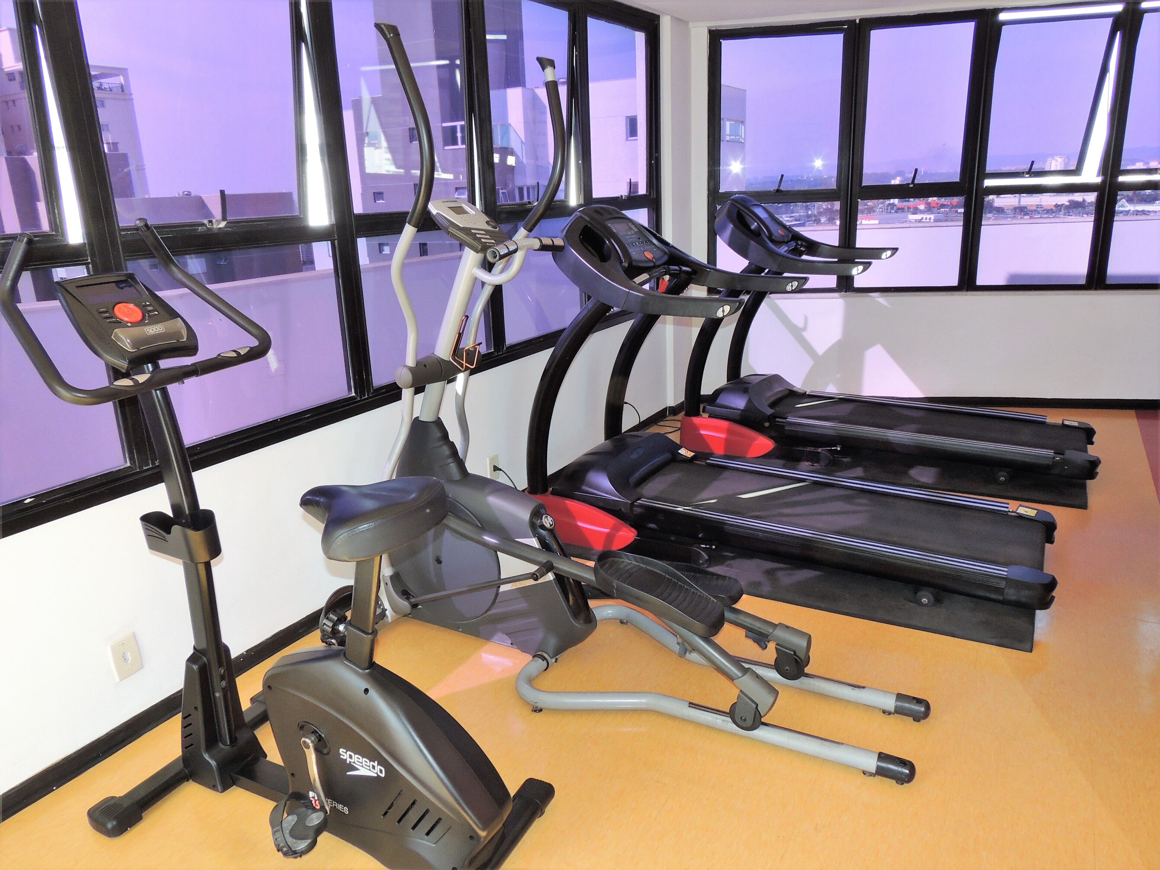 Un gimnasio con equipo de ejercicio, incluyendo cintas de correr y una máquina elíptica, amplias ventanas con vistas a edificios y suelo de madera.