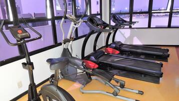 Un gimnasio con equipo de ejercicio, incluyendo cintas de correr y una máquina elíptica, amplias ventanas con vistas a edificios y suelo de madera.