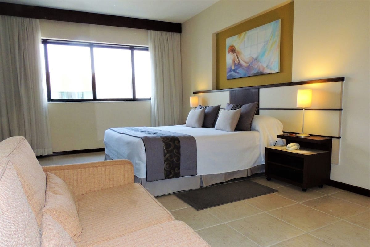 Habitación de hotel con cama, mesita de noche con lámpara, un cuadro en la pared y ventana con cortinas.