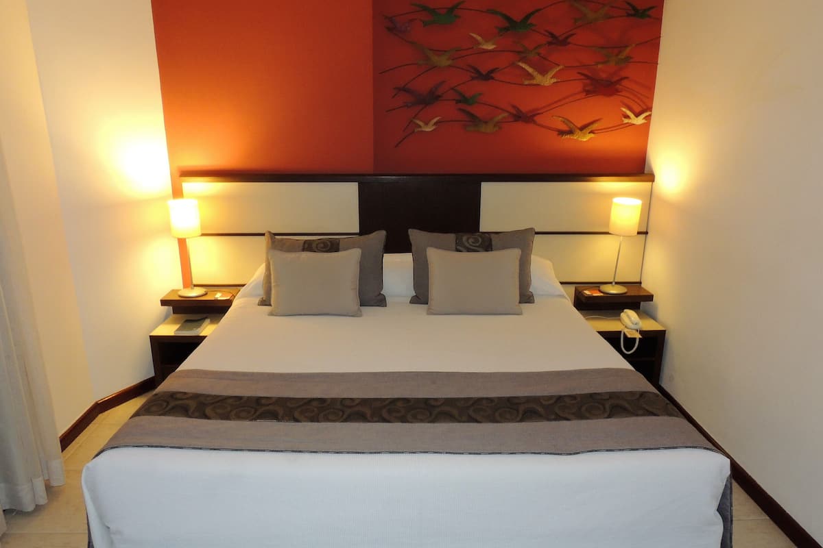 Habitación de hotel con una cama grande, dos mesitas de noche con lámparas, un teléfono y una pared con un mural de aves.