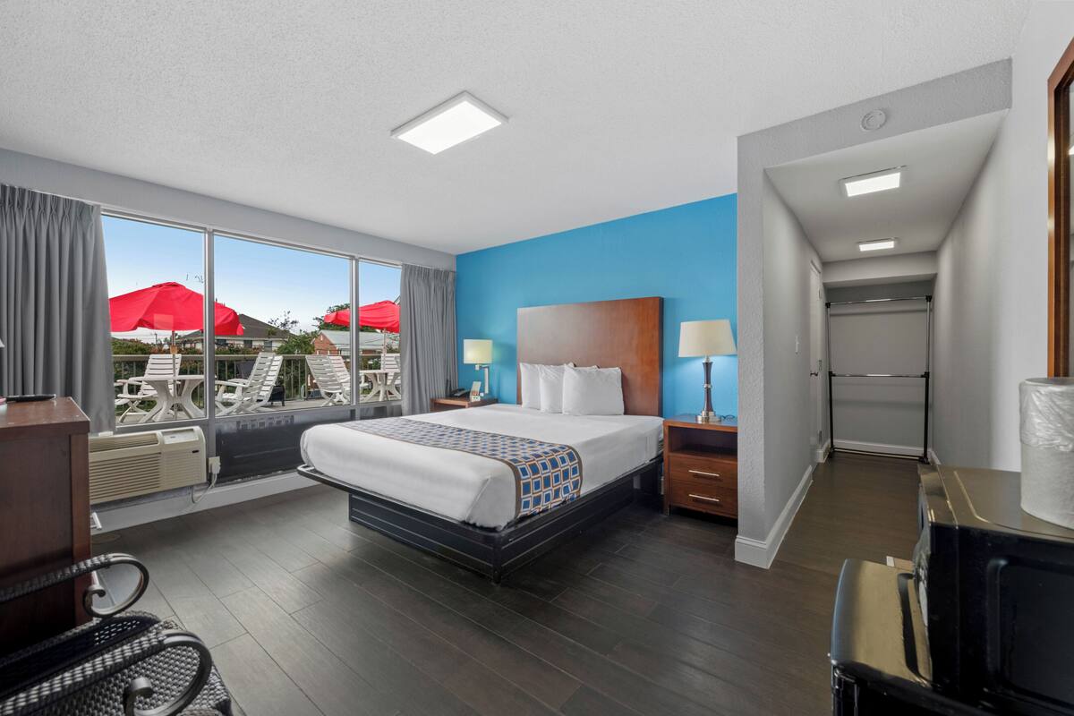 A hotel room with a large bed, a desk, a chair, and a view of outdoor seating with red umbrellas.