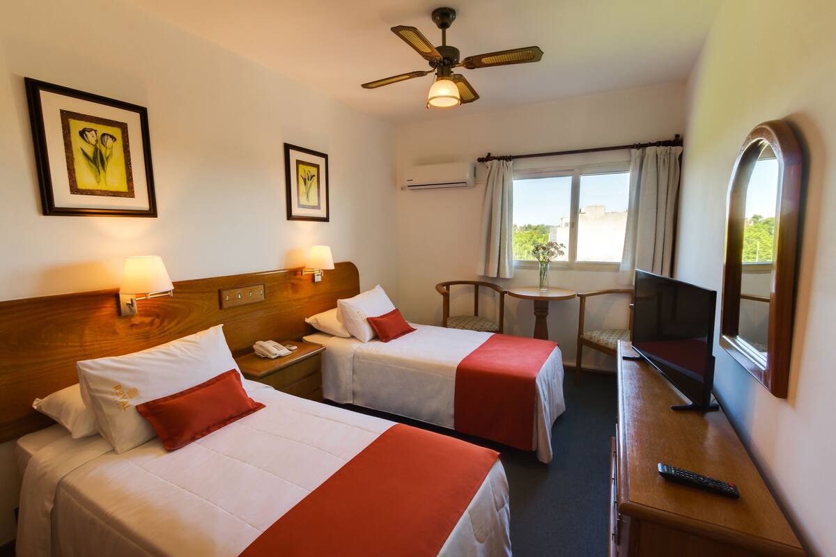 A hotel room with two beds, a desk, a chair, a TV, and a ceiling fan.