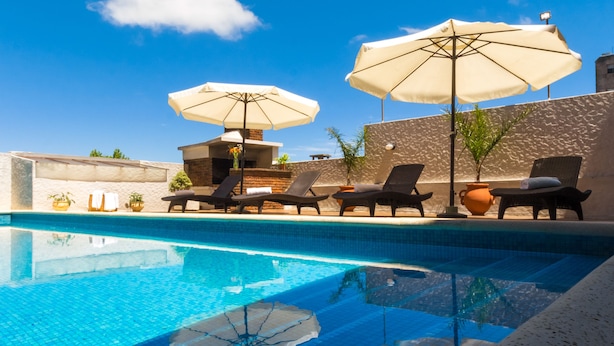 A pool area with lounge chairs and umbrellas, surrounded by a stone wall and plants. - Hotel Royal Colonia (Colonia del Sacramento)