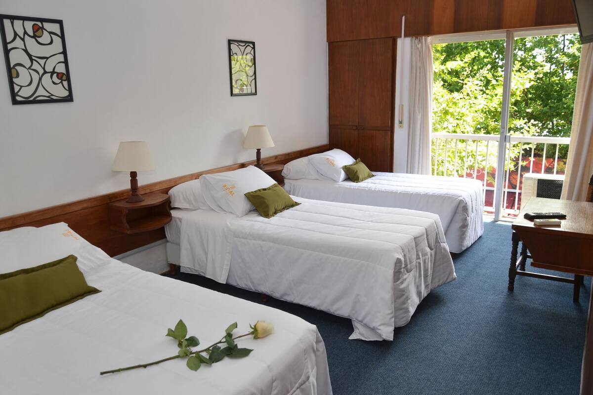 A hotel room with three single beds, a wooden headboard, a desk with a chair, and a balcony with a view of greenery.