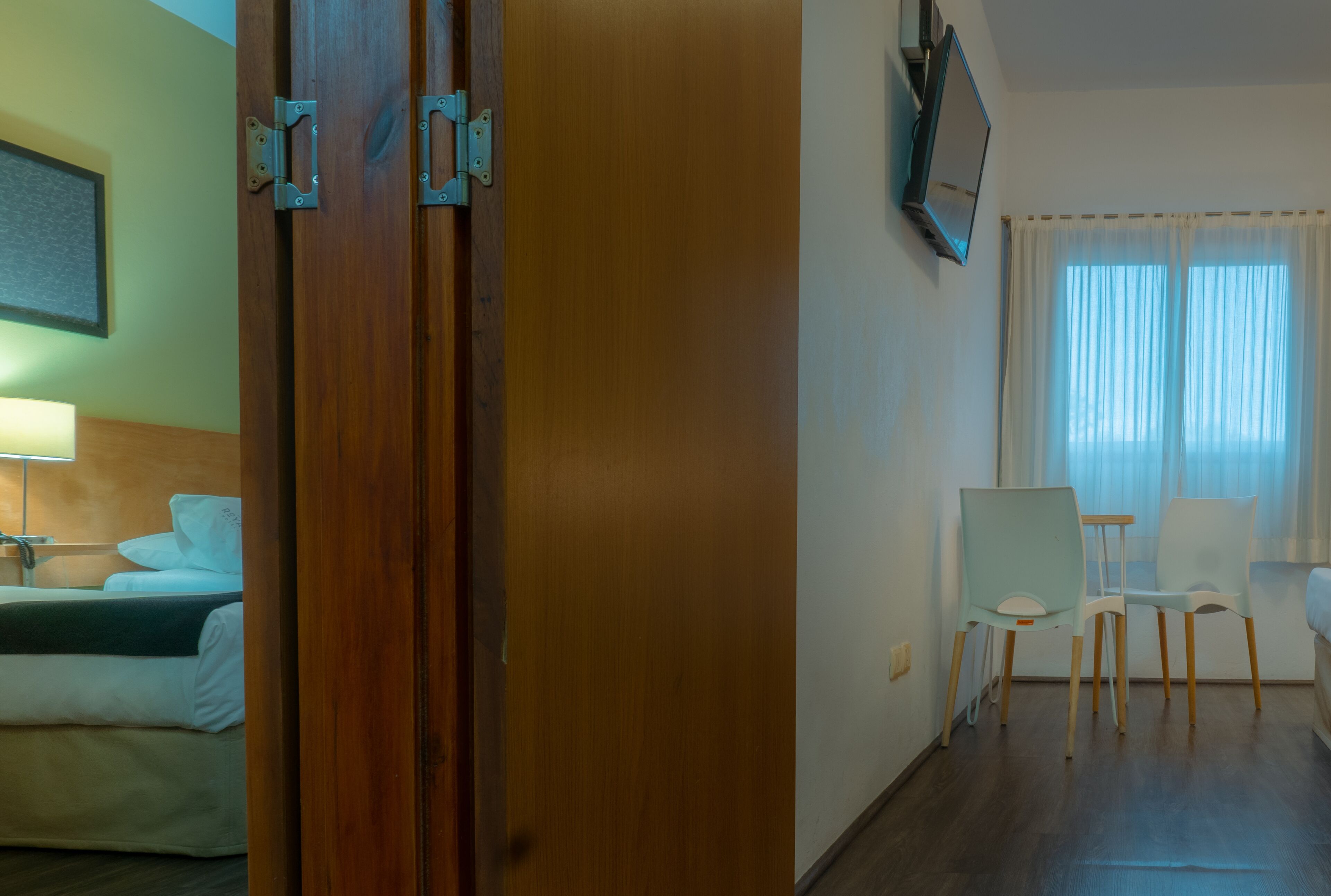 Habitación de hotel con puerta de madera, cama con ropa blanca, escritorio con silla y televisor montado en la pared.