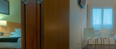 Habitación de hotel con puerta de madera, cama con ropa blanca, escritorio con silla y televisor montado en la pared.