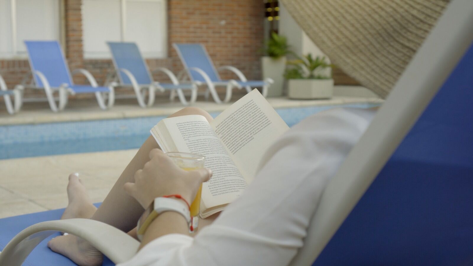 Una persona descansando en una tumbona junto a la piscina, leyendo un libro y sosteniendo un vaso de jugo de naranja.