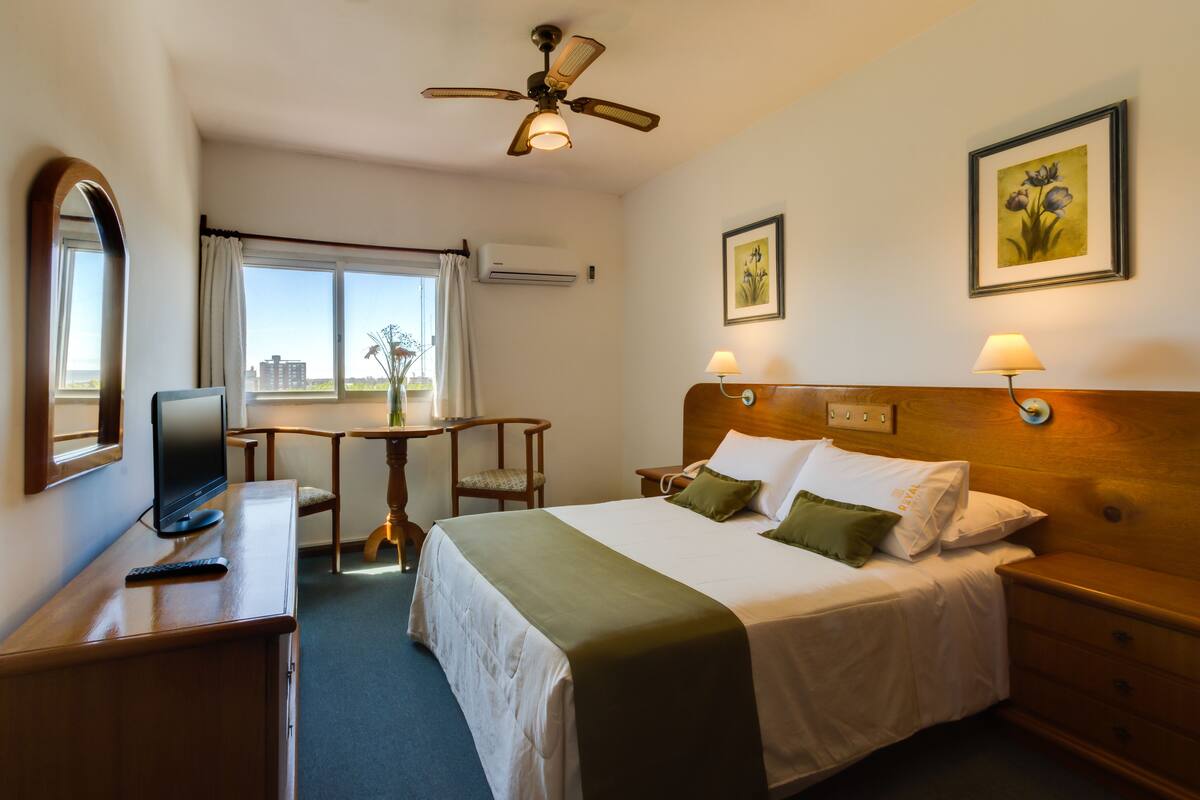A hotel room with a bed, bedside lamps, a desk with a computer, a chair, and a window with a view of buildings.