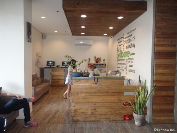 Un área de recepción con un mostrador de madera, un cartel de bienvenida en varios idiomas y algunas plantas en macetas.