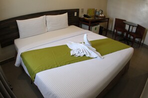 Un letto ben rifatto con lenzuola bianche e un dettaglio verde, una piccola scrivania con una sedia e un telefono fissato al muro.