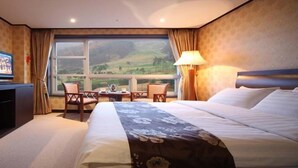 A hotel room with a large bed, a desk with a chair, a television, and a view of a green landscape through a large window. - Orient Hotel & Resort (Pyeongchang)