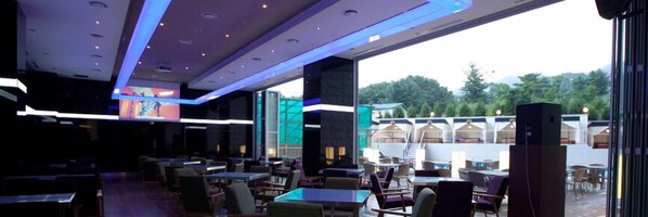 Un restaurante moderno con mesas dentro y fuera, ventanas amplias y vistas a zonas verdes.