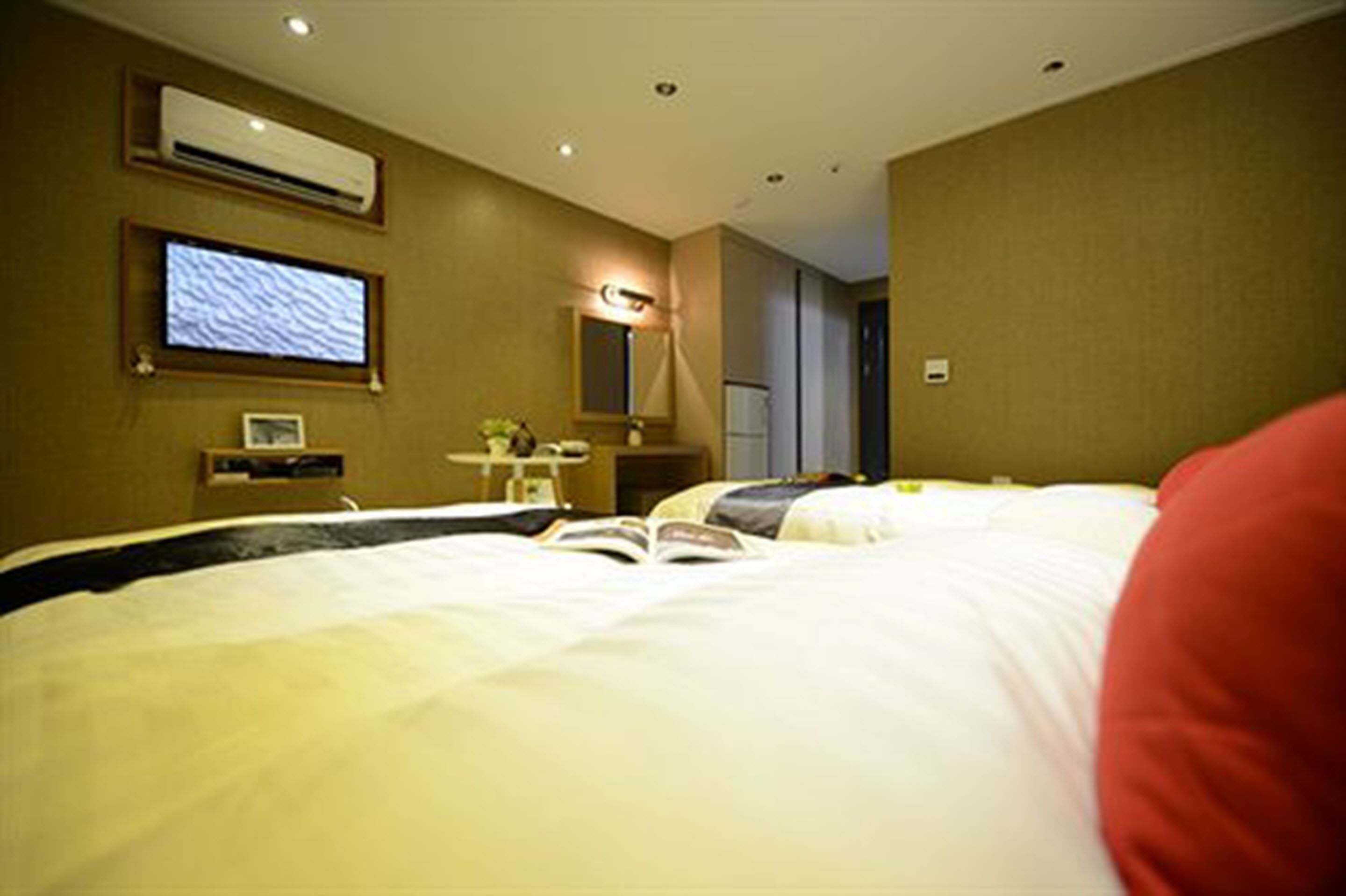 A hotel room with a bed, a desk with a computer, a TV, and an air conditioning unit on the wall.