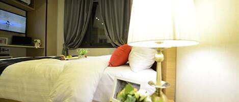 A hotel room with a bed, a bedside lamp, a TV on the wall, and a window with curtains.