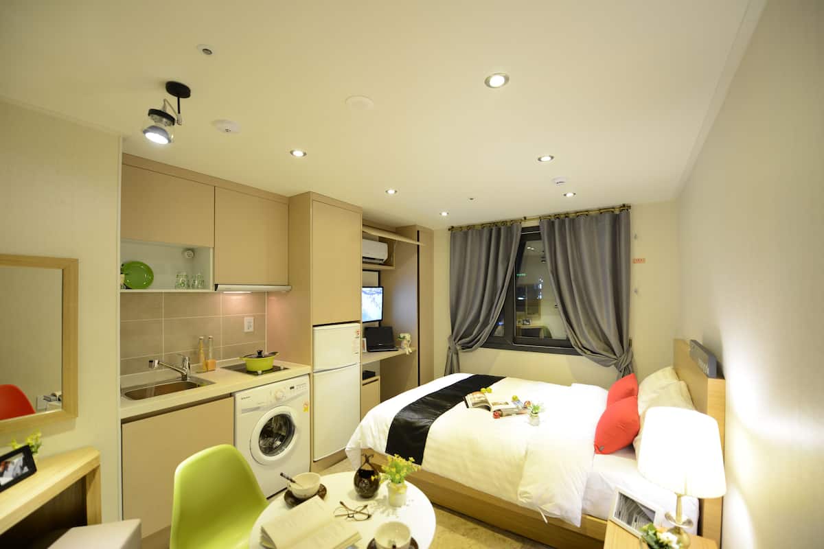 A compact hotel room with a kitchenette, a bed with a black and white bedspread, a bedside table with a lamp, and a window with curtains.