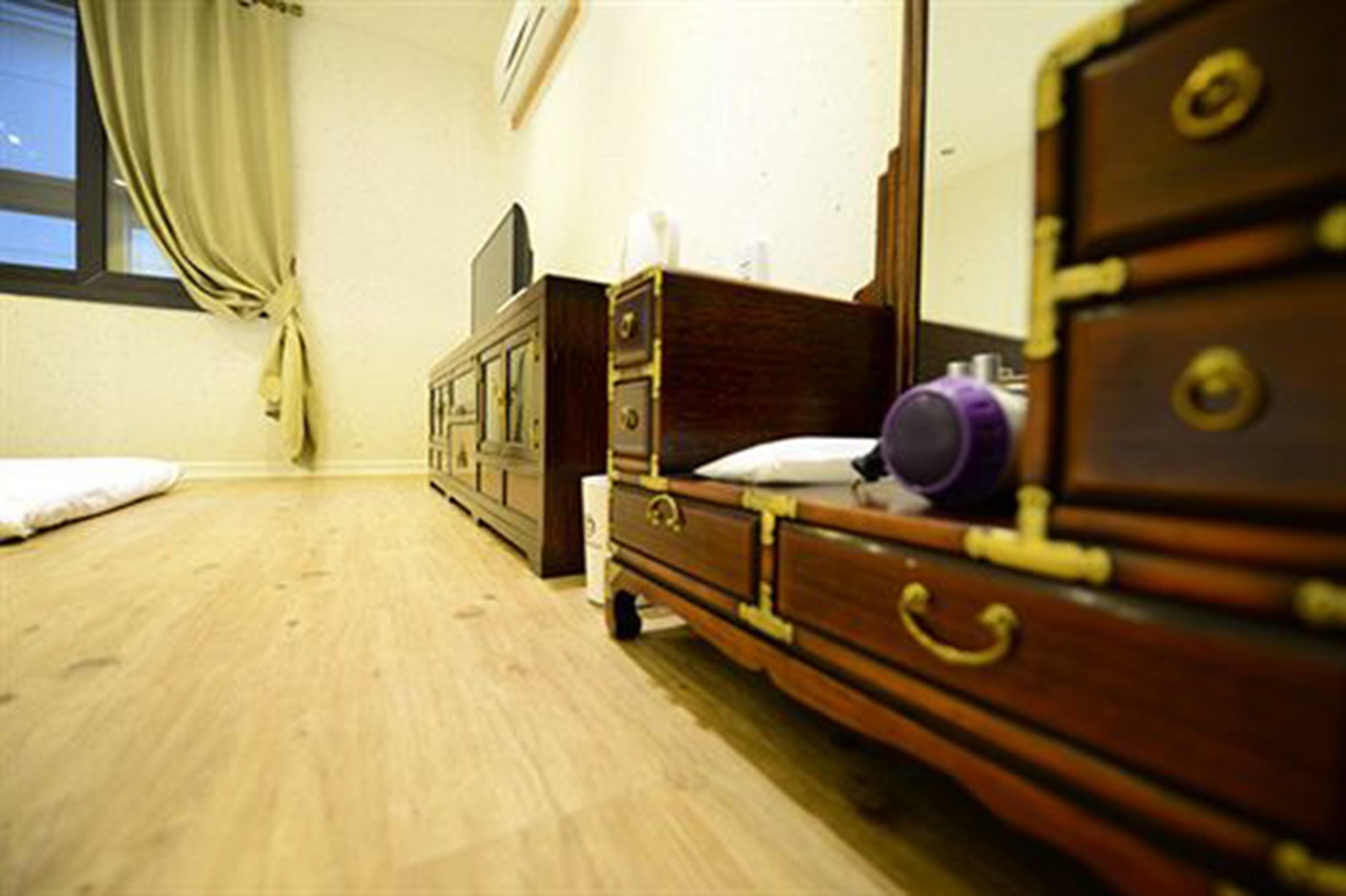 Une chambre avec une commode en bois, un lit avec un oreiller blanc, un téléviseur posé sur un meuble et une fenêtre avec des rideaux.