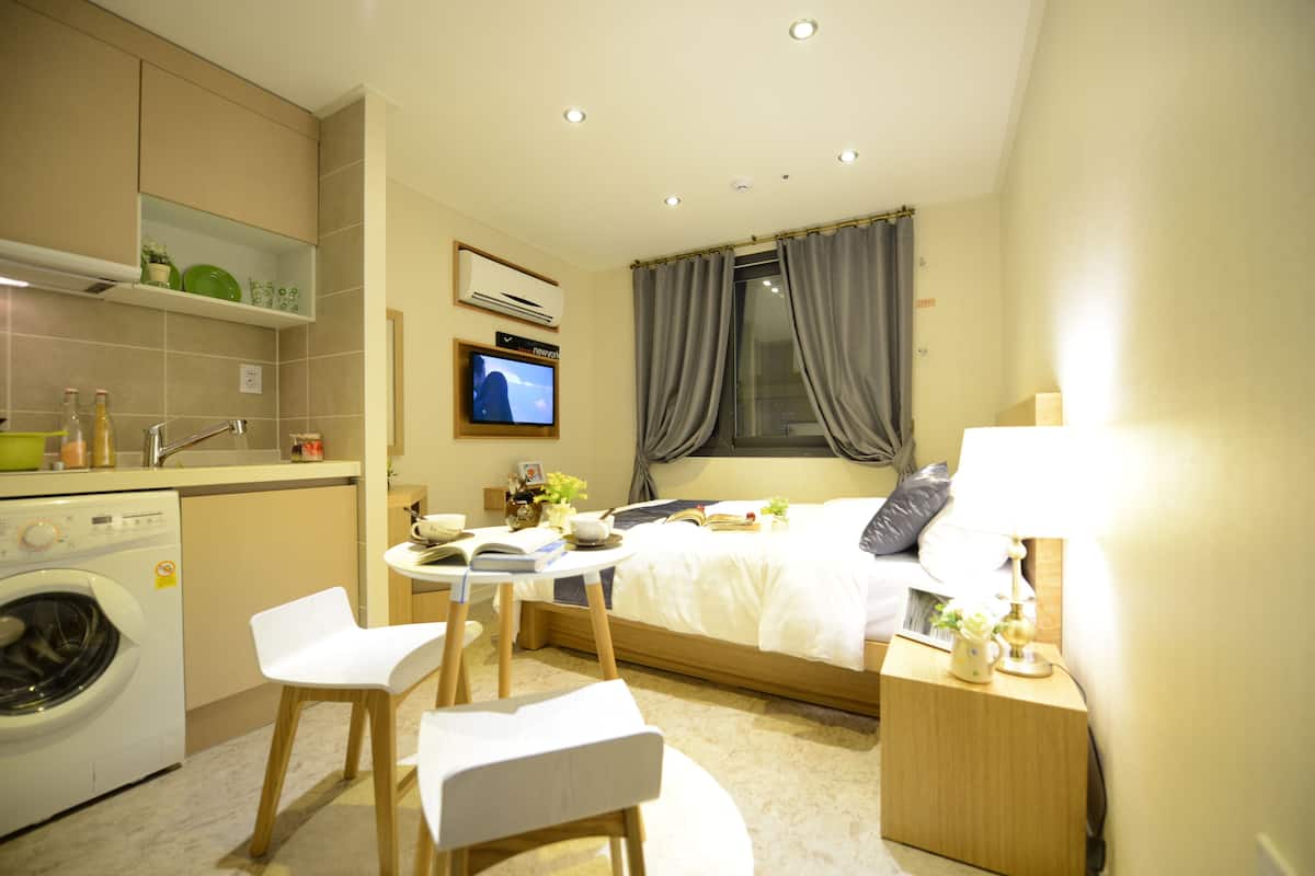A compact hotel room with a bed, bedside table, TV, and a small kitchenette with a washing machine.