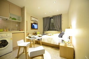 Ein kompaktes Hotelzimmer mit Bett, Nachttisch, Fernseher und einer kleinen Küchenzeile inklusive Waschmaschine.