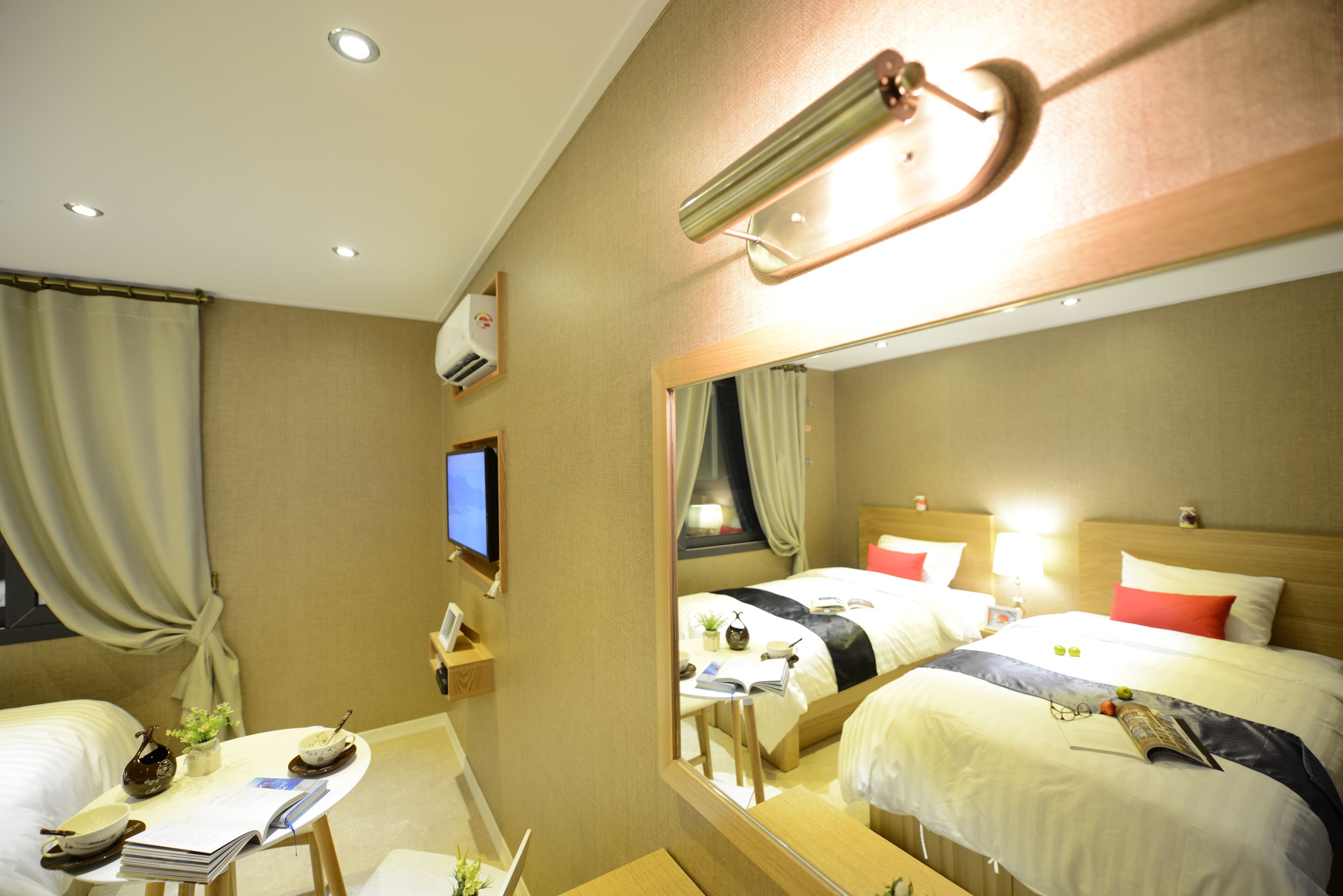 A hotel room with two beds, a desk with a chair, a small table with a plant, a mirror, and a television on the wall.