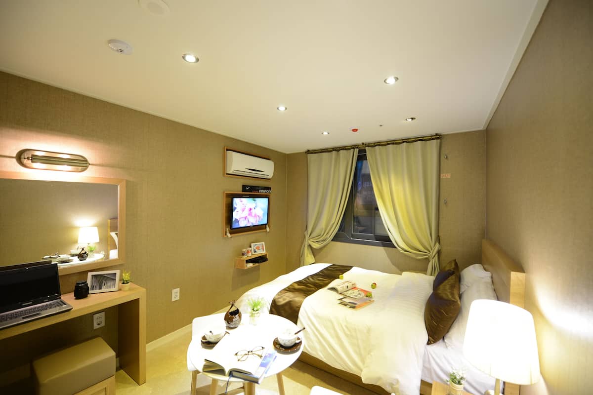 A hotel room with a bed, a desk with a laptop, a television, and a small table with a cup and a plant.