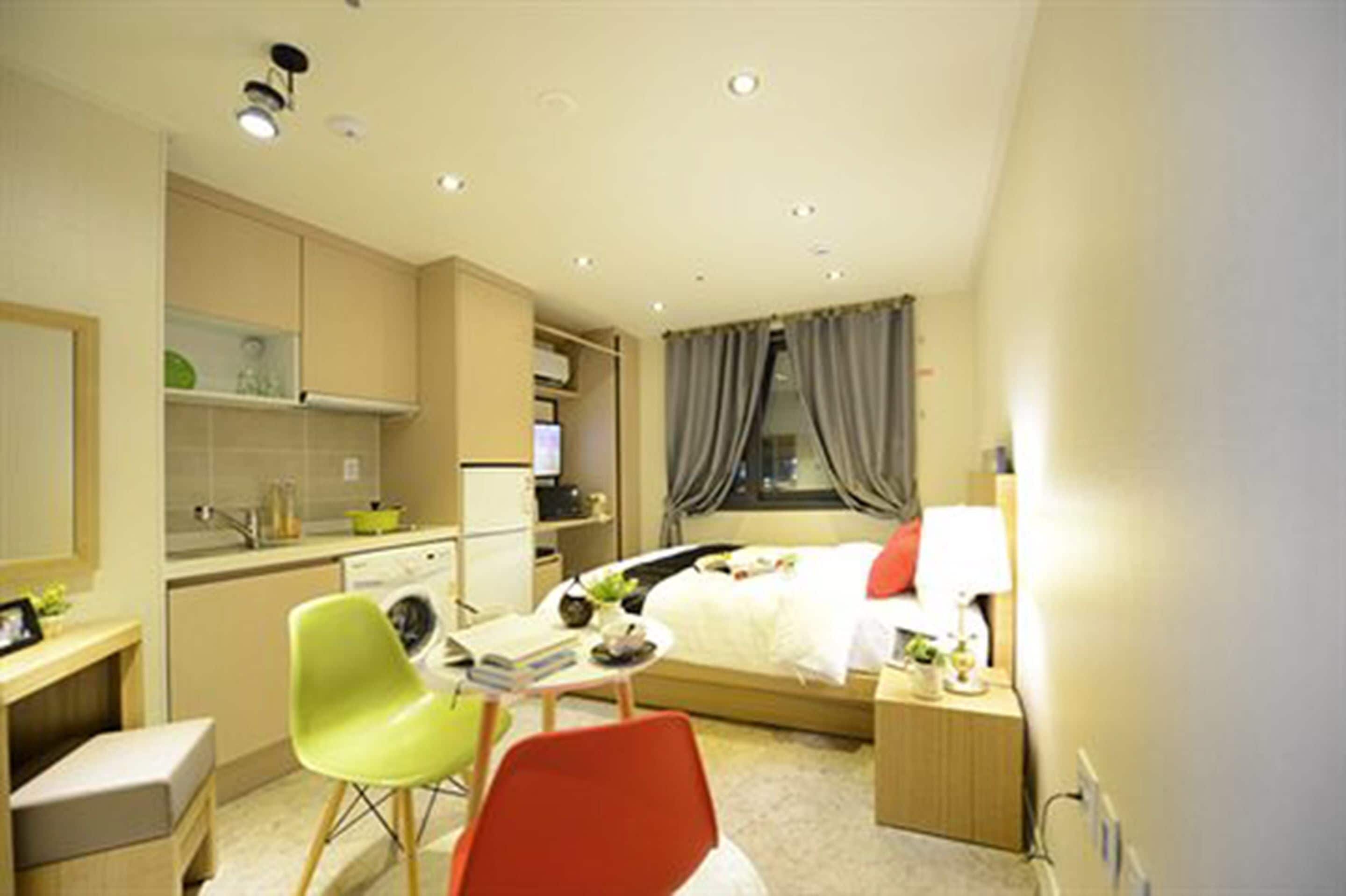 Una habitación de hotel con cama, escritorio, silla, lavadora y una pequeña cocina.