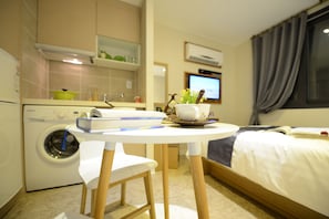Ein kleines Hotelzimmer mit Waschmaschine, einem kleinen Esstisch mit Stuhl, einem Bett mit Fernseher und einem Fenster mit Vorhängen.
