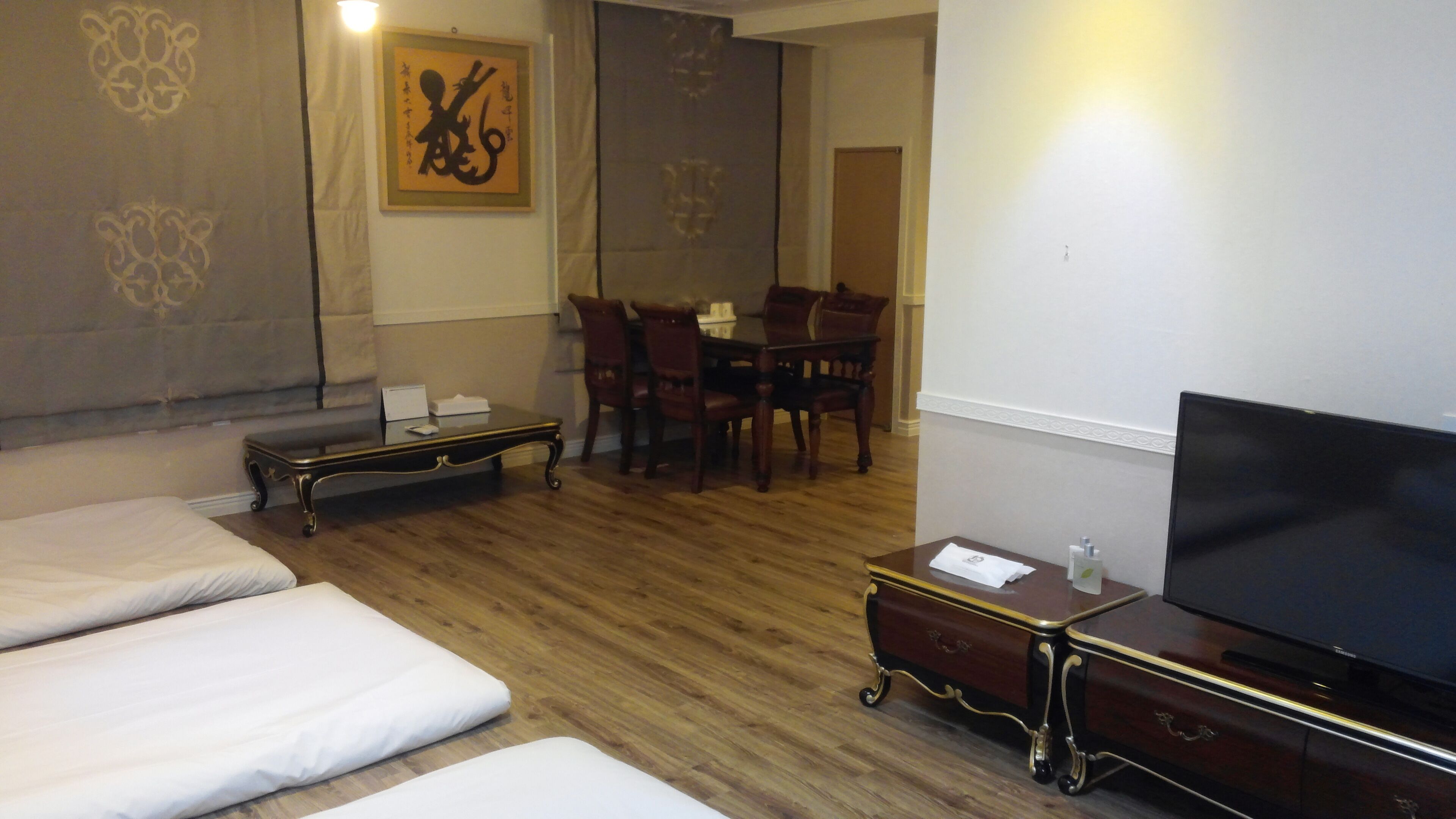 Una habitación de hotel con suelo de madera, televisión, una mesita con un control remoto, cama, mesa de comedor con sillas y un adorno en la pared.