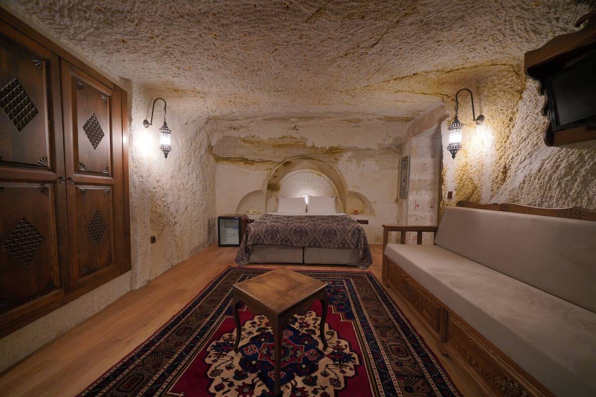 A room with a wooden floor, a bed with a patterned bedspread, a sofa, a small wooden table, and wall-mounted lights.