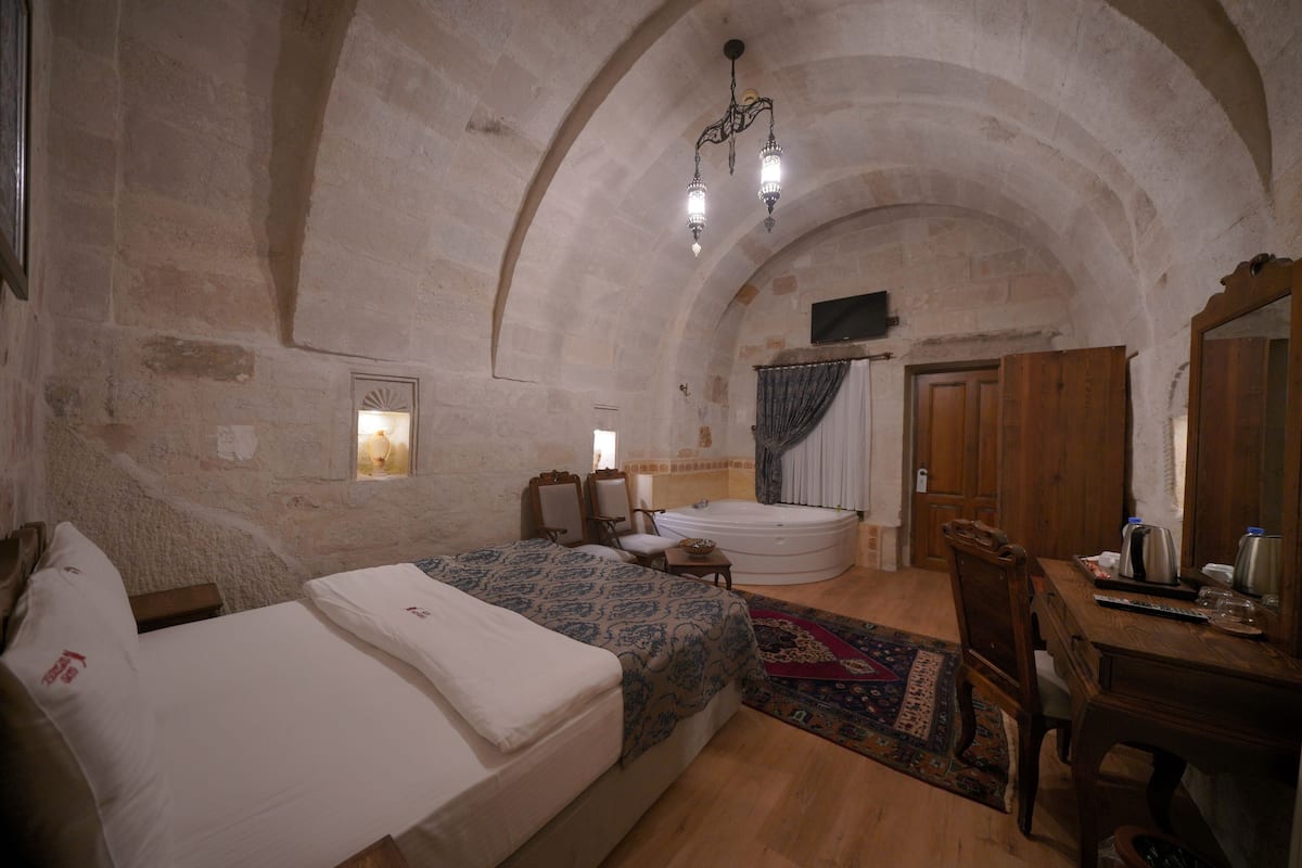 A bedroom with a large bed, a wooden desk, a chair, a small table, a bathtub, and a chandelier.