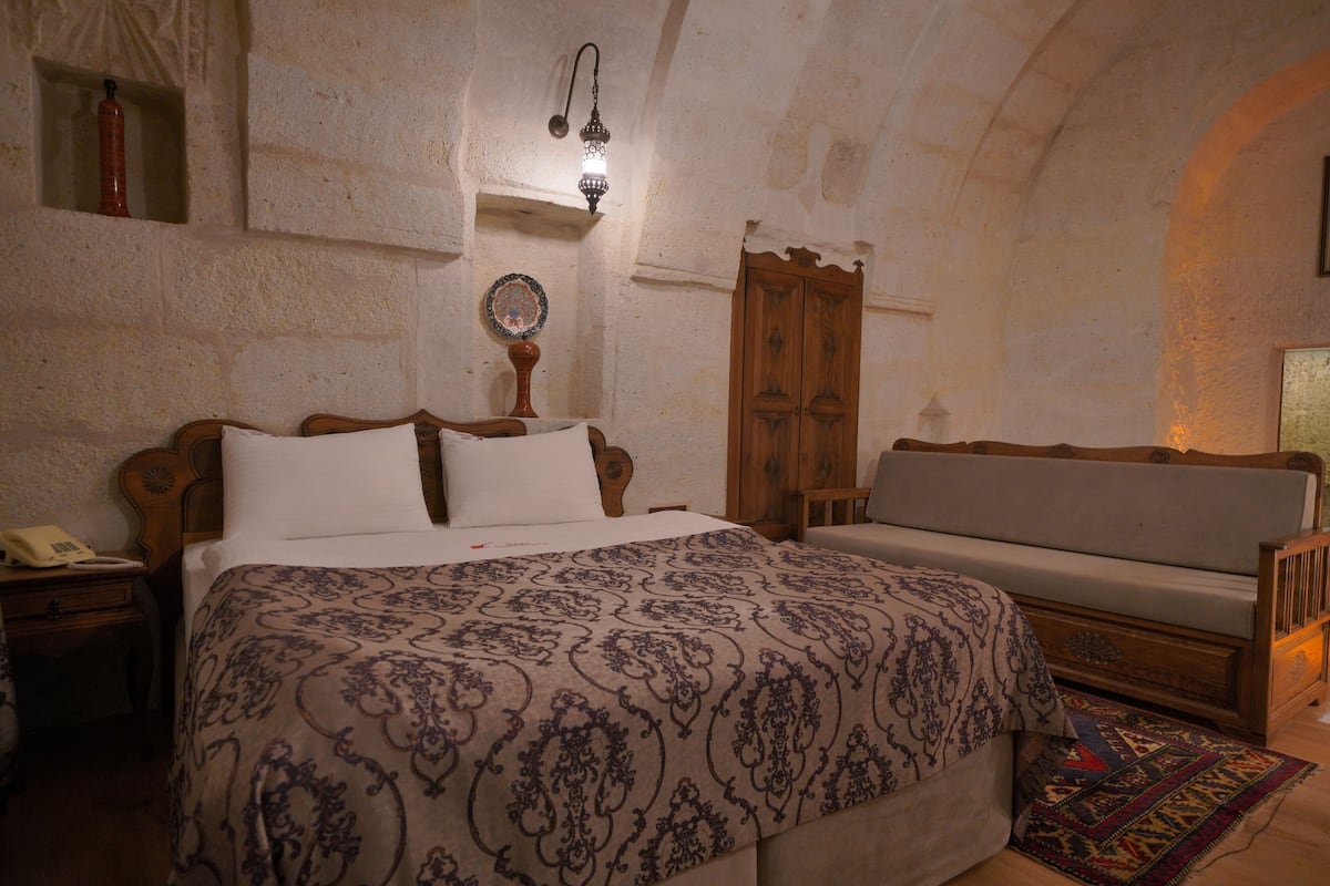 A bedroom with a large bed, a wooden wardrobe, a bedside table with a lamp, and a small shelf with a decorative item.