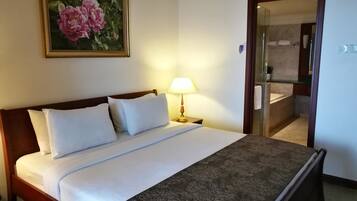 A hotel room with a bed, a bedside lamp, a painting of flowers, and a bathroom visible through an open door.