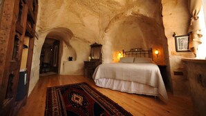 Un dormitorio rústico con piso de madera, cama con cubrecama blanco, mesita de noche con lámpara y pared de piedra.