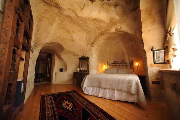 A rustic bedroom with a wooden floor, a bed with a white bedspread, a nightstand with a lamp, and a stone wall. - Anitya Cave House (Ürgüp)