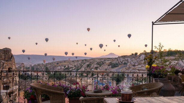 Una terraza con mobiliario de mimbre, una mesa con una taza y vistas de globos aerostáticos sobre un paisaje montañoso.