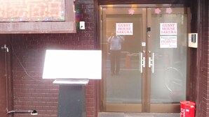 A guest house entrance with two doors labeled "GUEST HOUSE SAKURA," a digital display, and a red barrel. - SAKURA GUEST HOUSE OSAKA DOUTONBORI - Hostel (Osaka)