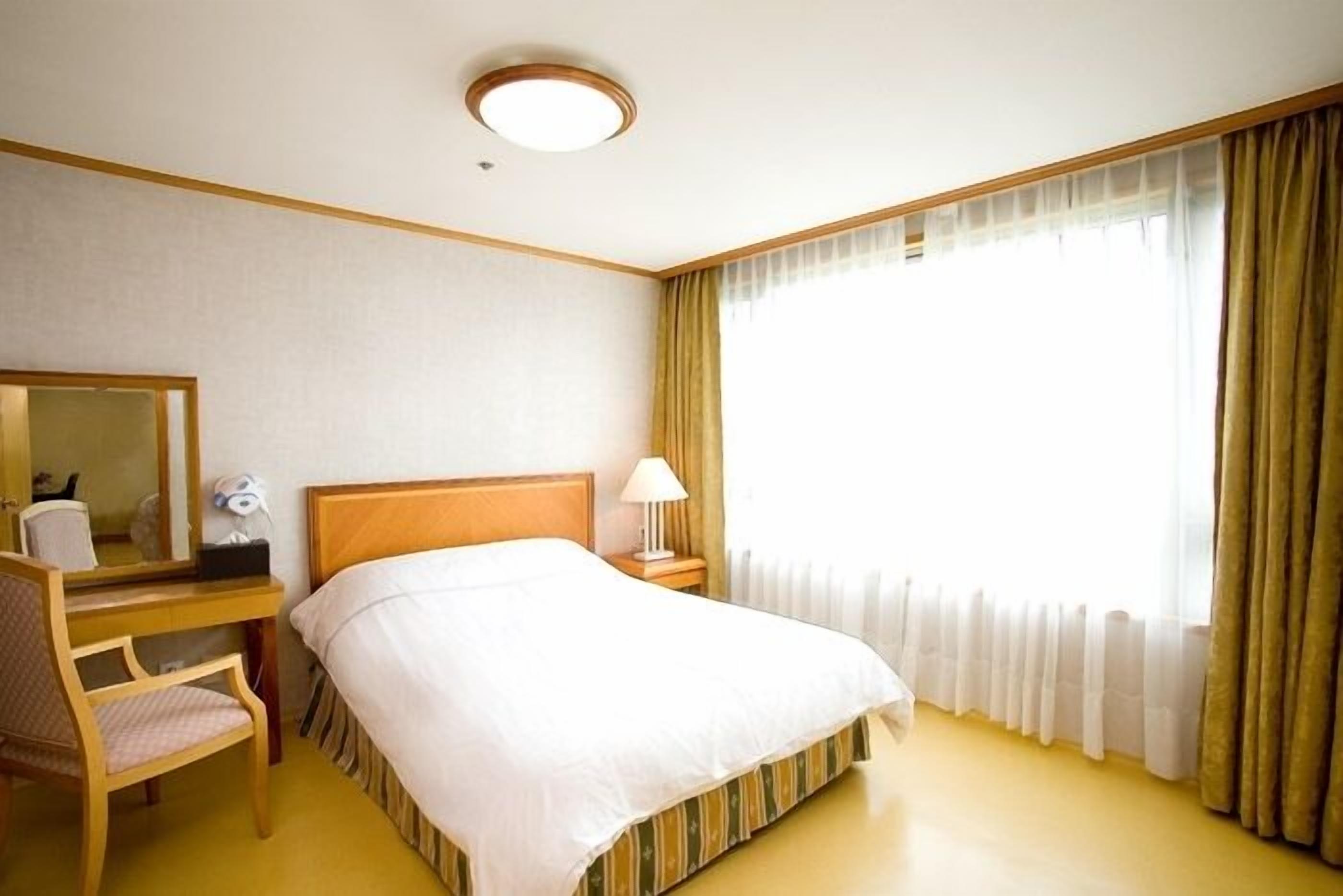 Et hotelværelse med en seng, et skrivebord med stol, et spejl, en lampe og et vindue med gardiner.