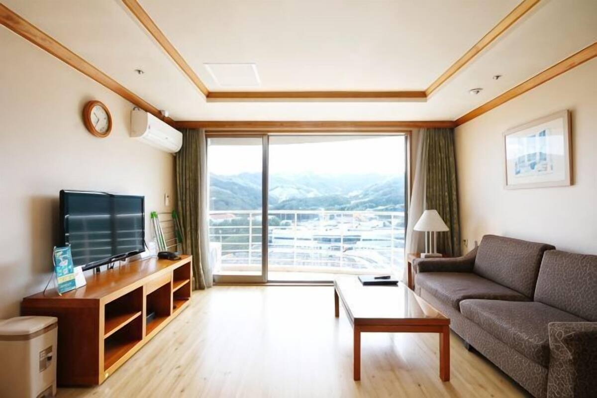 Un salon spacieux doté d’une grande fenêtre, d’un téléviseur à écran plat, d’un canapé, d’une table basse et d’un meuble de rangement en bois pour les appareils multimédias.