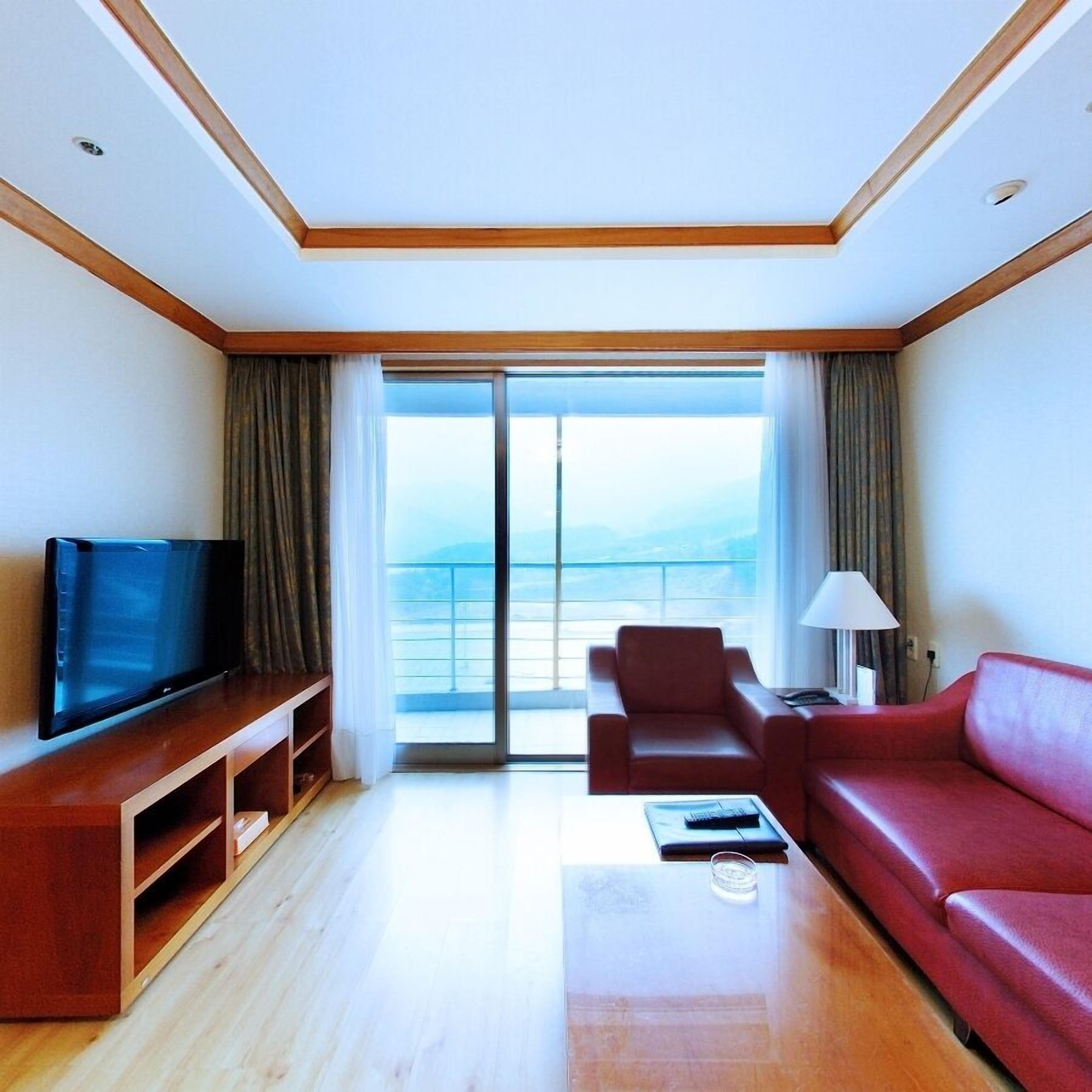 A hotel room with a large window, a television, a red sofa, a coffee table, and a lamp.