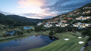 Un campo de golf con un estanque, rodeado de edificios modernos de varios pisos y amplias ventanas, con montañas y un cielo al atardecer al fondo.