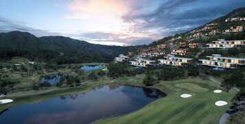 A golf course with a pond, surrounded by modern multi-story buildings with large windows, set against a backdrop of mountains and a sunset sky.
