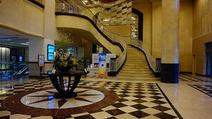 Un vestíbulo con piso a cuadros, un arreglo floral central, una escalera curva y un quiosco informativo digital.