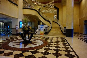 A lobby with a checkered floor, a central floral arrangement, a curved staircase, and a digital information kiosk.
