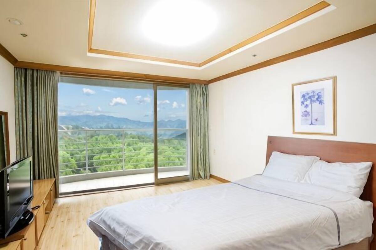 Une chambre à coucher avec un lit, une télévision, une grande fenêtre donnant sur un paysage verdoyant et montagneux, et un tableau encadré accroché au mur.
