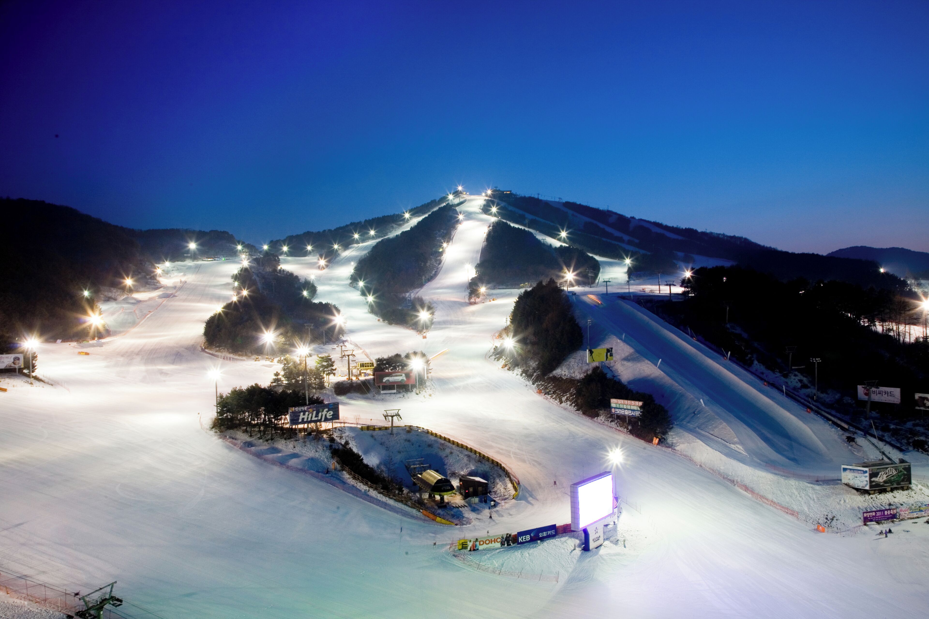 A snow-covered ski slope at night with artificial lighting and signage.