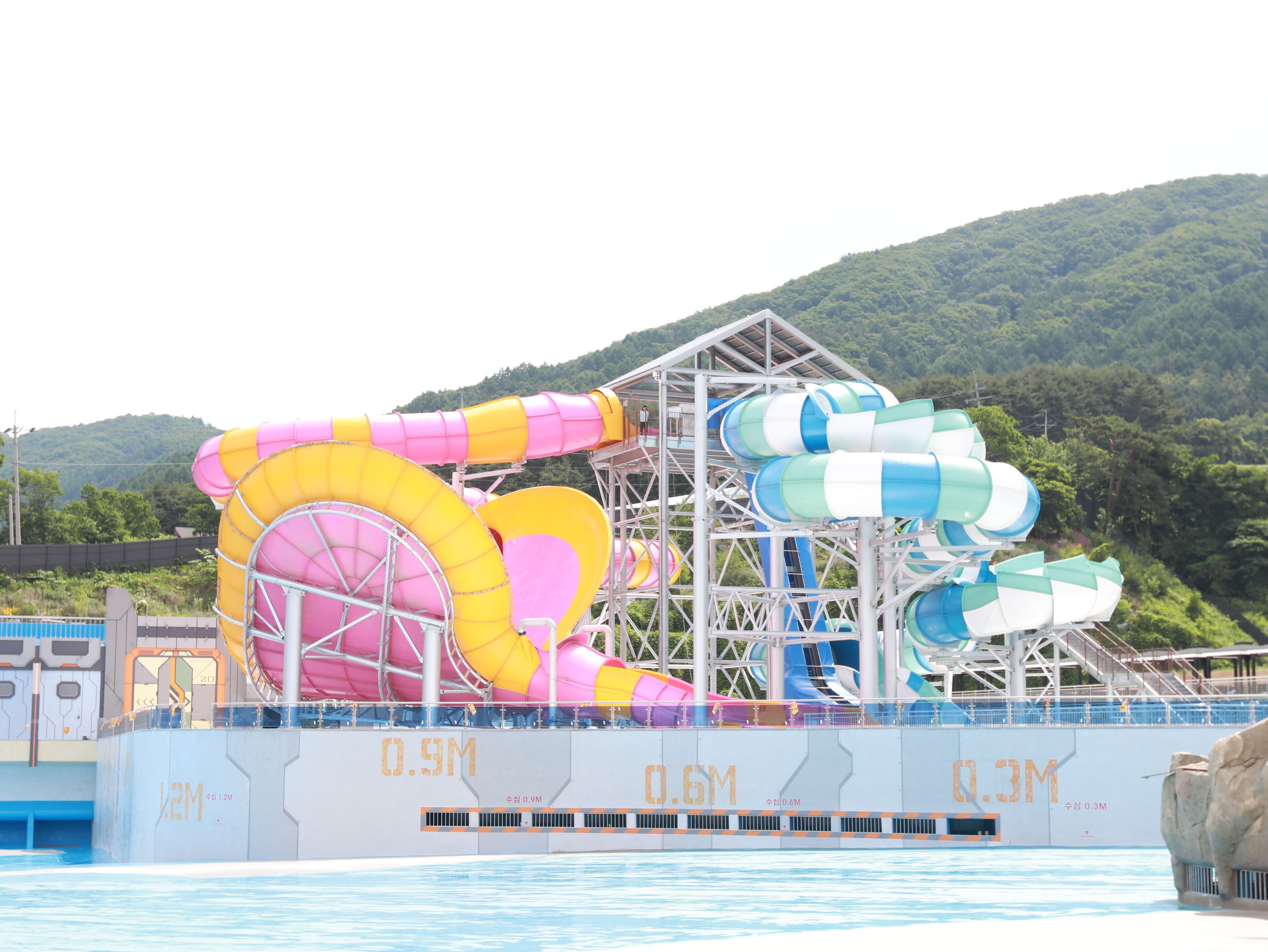A water park with multiple water slides, including a large pink and yellow slide and a blue and white slide. The park is surrounded by a metal structure and has a pool in the foreground.