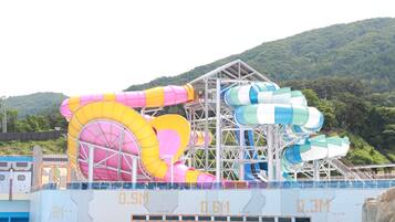 A water park with multiple water slides, including a large pink and yellow slide and a blue and white slide. The park is surrounded by a metal structure and has a pool in the foreground.