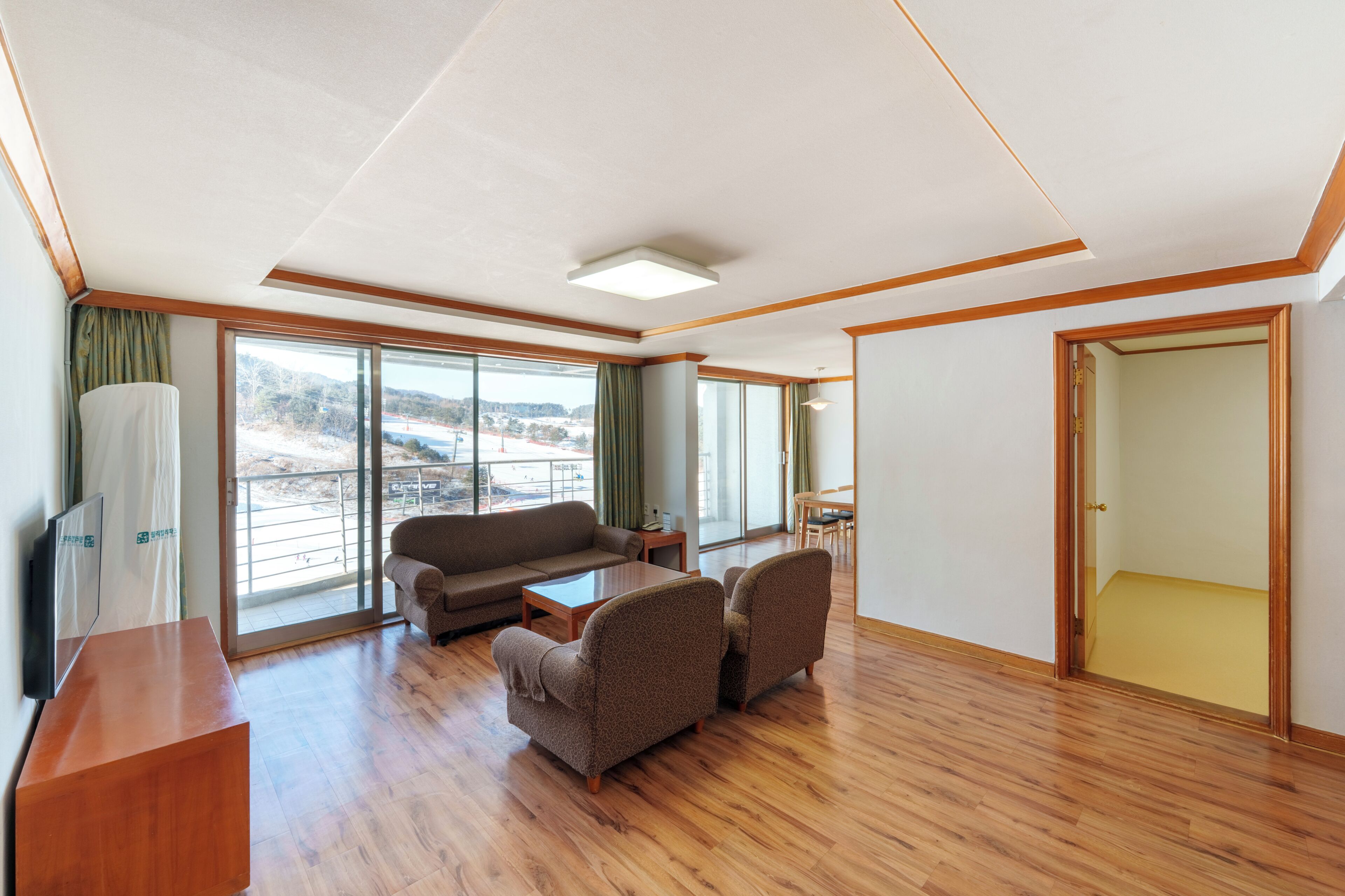 A spacious room with a wooden floor, a sofa, two armchairs, a small table, and a large window offering a view of a snowy landscape.