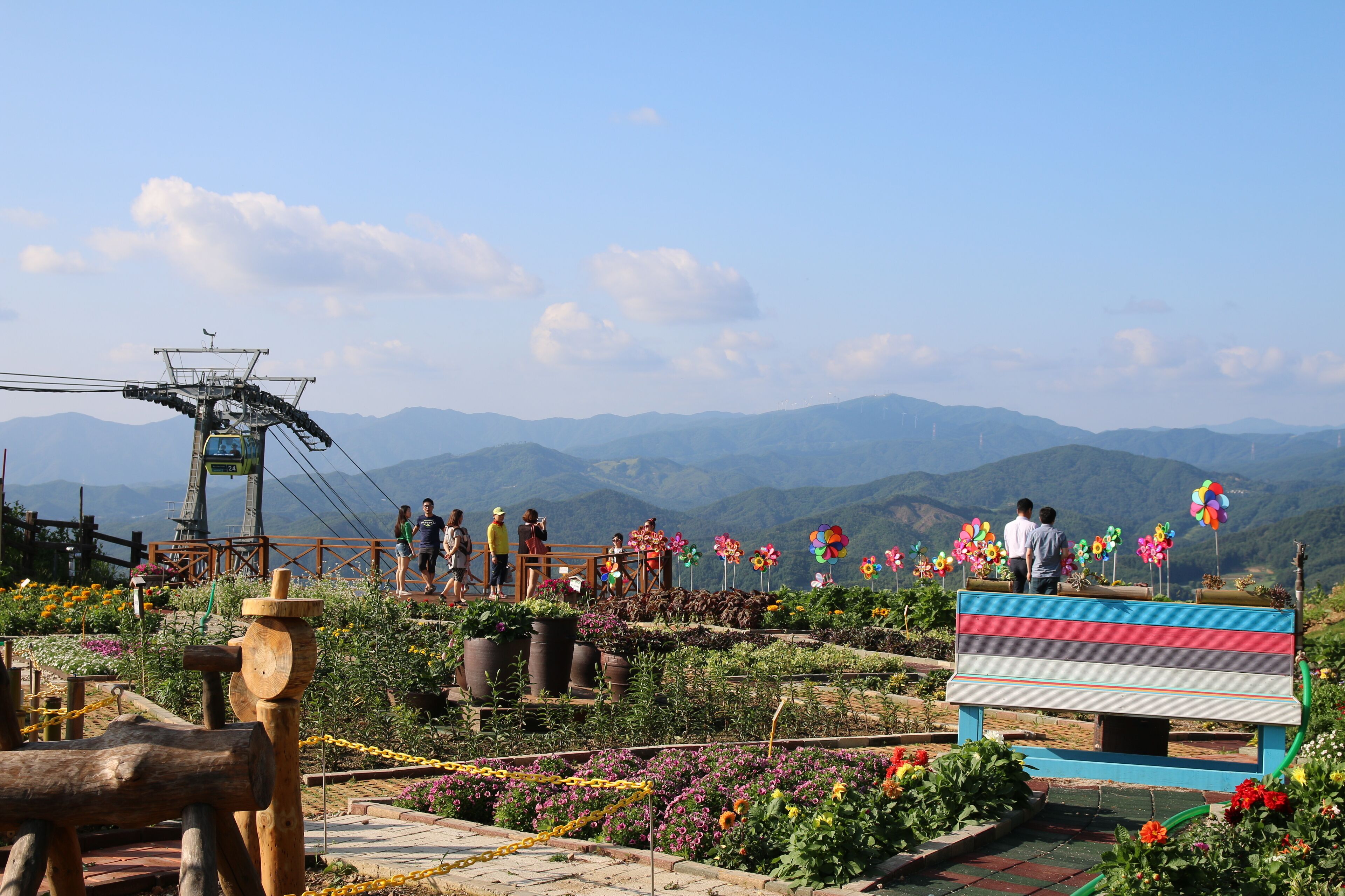 Un área al aire libre con un paisaje hermoso, un banco de madera, macizos de flores y la estructura de un teleférico.