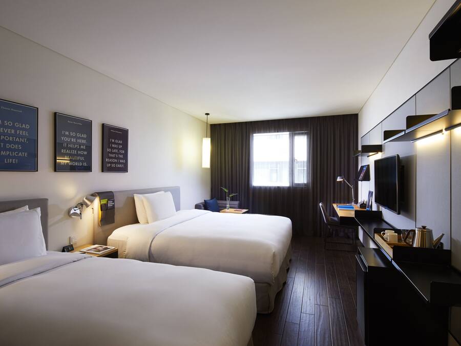 Quarto de hotel com duas camas, uma escrivaninha, televisão de tela plana e janela com cortinas.